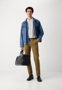 Denimjakke, lys grå polo, khaki bukser, sort læder duffeltaske, sorte snøresko; model står i en neutral indstilling.