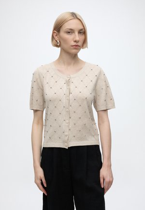 Femme aux cheveux blonds courts porte un cardigan beige à manches courtes boutonné avec de petites perles métalliques et un pantalon noir sur un fond uni.