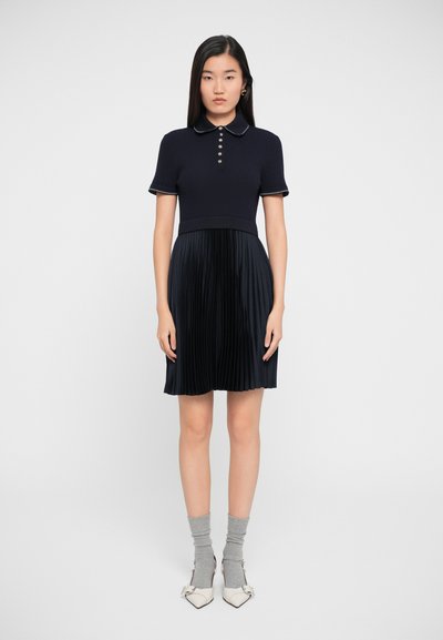 Abito polo blu navy con top a coste e gonna plissettata, dotato di bottoni e colletto a contrasto. Abbinato a calze grigie e scarpe con tacco bianche.