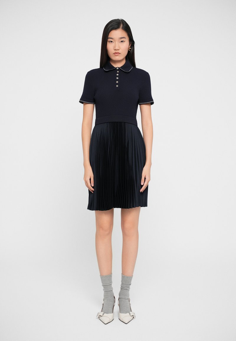 Navy polo-mekko, jossa on ribattu yläosa ja laskostettu hame, varustettuna nappeilla ja kontrastivärisellä kauluksella. Tyylitelty harmailla sukilla ja valkoisilla korkokenkillä.