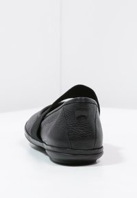 Zapato de cuero negro con tira elástica, puntera redonda y superficie texturizada. Diseño minimalista sin marcas visibles ni detalles de herrajes.