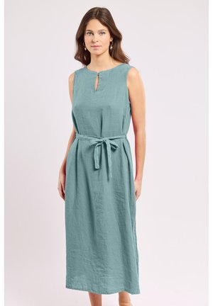 Femme aux cheveux châtain moyen portant une robe midi sans manches de couleur teal clair avec ceinture à la taille et petite encolure goutte en avant, debout devant un fond uni.