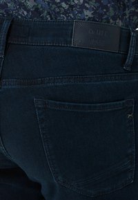 Dark blue denim jeans with back pocket and black leather patch labeled "Hi-FLEX BRAX" worn with a blue patterned shirt.