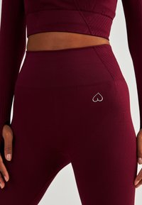 Bordowe legginsy z wysokim stanem, wykonane z teksturowanej tkaniny, z małym srebrnym logo w kształcie serca na plecach, zaprojektowane do użytku sportowego.