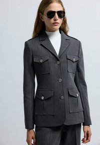 Chaqueta gris ajustada con cuatro bolsillos de parche, acentos de botones plateados y un diseño estructurado. Llevada sobre una blusa de cuello alto.