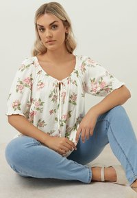 Vit blus med blommor i rosa och gröna blad, med rundad halsringning och rynkade ärmar. Bärs med ljusblå jeans.