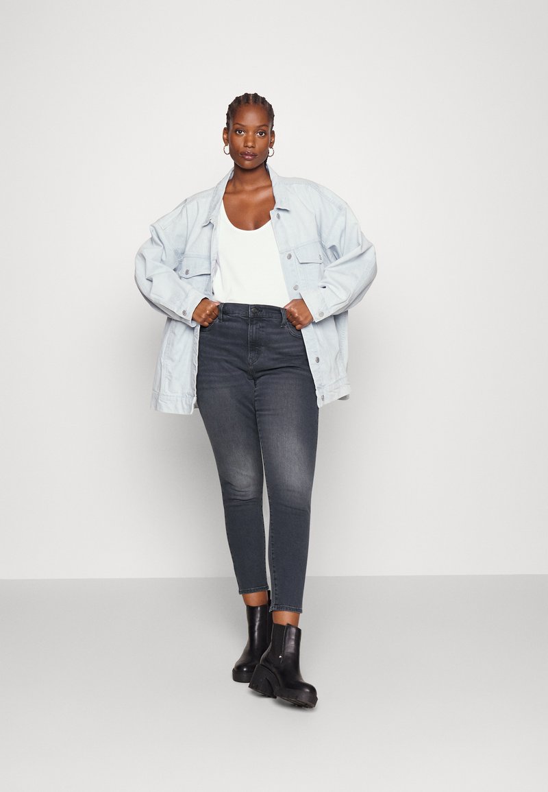 Hellblaue, übergroße Jeansjacke über einem weißen Tanktop, kombiniert mit engen, dunklen Jeans und schwarzen Stiefeletten. Sanfte Texturen und lässige Passform.