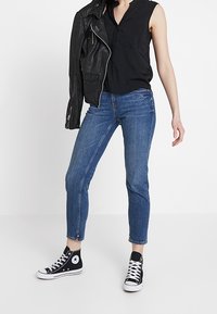 Blouse noire sans manches avec une patte de boutonnage frontale, associée à un jean denim bleu et des baskets montantes noires, portant une veste en cuir noire.