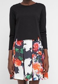 Svart långärmad tröja i kombination med en blommig kjol med orange, rosa och gröna mönster på en vit bakgrund. Texturerad tyg.