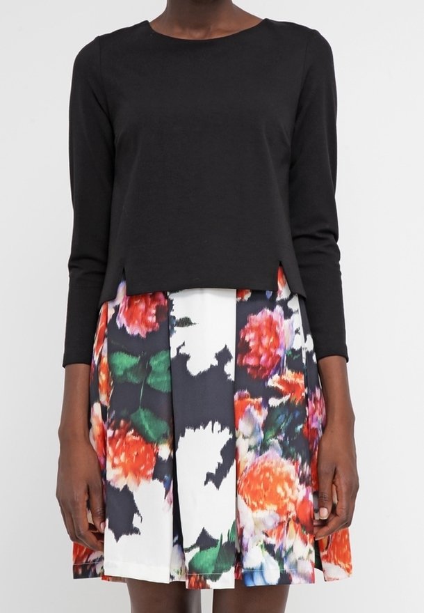 Svart långärmad tröja i kombination med en blommig kjol med orange, rosa och gröna mönster på en vit bakgrund. Texturerad tyg.