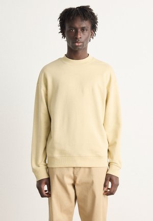 Šviesiai geltonas crewneck tipo megztinis, pagamintas iš minkštos medžiagos, su gabo rankovėmis ir apykakle, turintis diskretišką logotipą ant krūtinės. Dėvimi su bežinėmis kelnėmis.