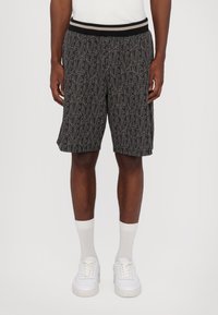Pantalones cortos negros con un patrón geométrico blanco, cinturilla elástica con rayas y un ajuste relajado que llega hasta la mitad del muslo, combinados con zapatillas blancas.