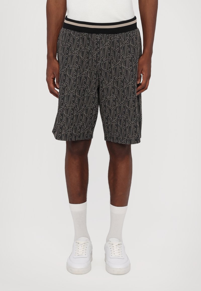 Pantalones cortos negros con un patrón geométrico blanco, cinturilla elástica con rayas y un ajuste relajado que llega hasta la mitad del muslo, combinados con zapatillas blancas.