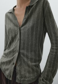 Person wearing a gray buttoned cardigan with thin white vertical stripes and dark pants, with one hand resting on their hip.