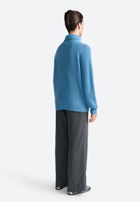 Pulover ribat albastru deschis cu guler înalt, purtat împreună cu pantaloni gri lejeri. Are mâneci lungi și un croi lejer.