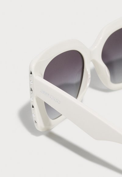 Lunettes de soleil blanches de forme carrée avec des verres teintés foncés et de petits clous décoratifs le long du bord extérieur de la monture.