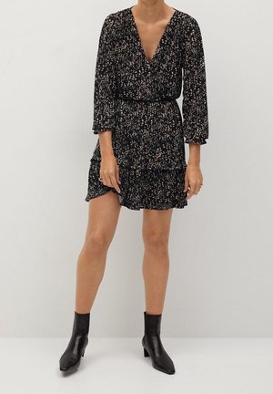 Femme portant une robe noire à manches longues avec un motif floral, un col en V et un ourlet à volants, associée à des bottines noires, sur un fond blanc uni.