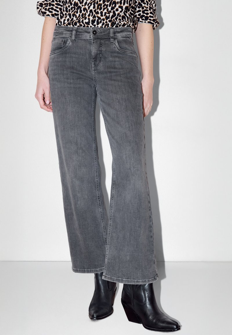 Personne portant un jean gris à jambes larges, des bottes noires à talons et un haut imprimé léopard, se tenant devant un fond blanc.