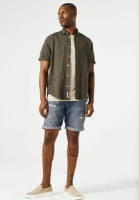 Camisa de botones de manga corta en verde oliva, camiseta gris claro debajo, pantalones cortos de mezclilla desgastados y zapatos slip-on beige.