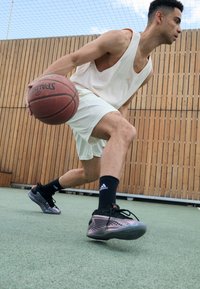Basketballspiller som dribler på en bane, iført en hvit mesh-topp, hvite shorts og svarte sokker med en Adidas-logo, samt svarte og rosa sneakers.