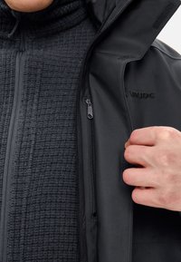 Giacca nera impermeabile con una fodera interna texturizzata, tasche con zip e colletto alto. Un branding discreto è visibile sul tessuto esterno.