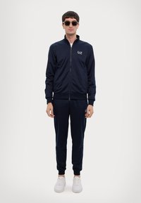 Costum sport marin format dintr-o jachetă cu fermoar și pantaloni asortați, având un logo mic. Asortat cu adidași albi și ochelari de soare negri. Textură netedă.