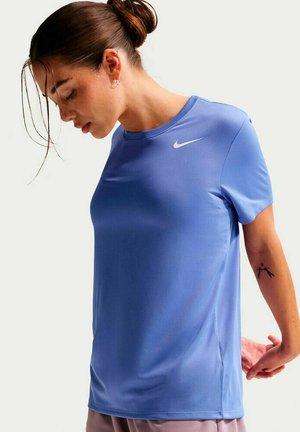 Mujer con cabello castaño recogido en un moño, vistiendo una camiseta azul de Nike, estirando los brazos detrás de la espalda, mirando hacia abajo.