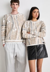 Pulovere tricotate cu model maro și bej, având manșete cu nervuri, purtate cu pantaloni plisati gri deschis și o fustă mini din piele neagră.