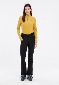 Maglia gialla in pile a maniche lunghe con colletto alto, abbinata a pantaloni da sci neri. Il modello indossa scarponi da sci neri, in piedi contro uno sfondo neutro.