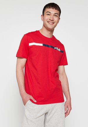 Homme souriant portant un t-shirt rouge FILA avec une bande blanche et marine, et un pantalon de survêtement gris clair, debout avec une main dans la poche.