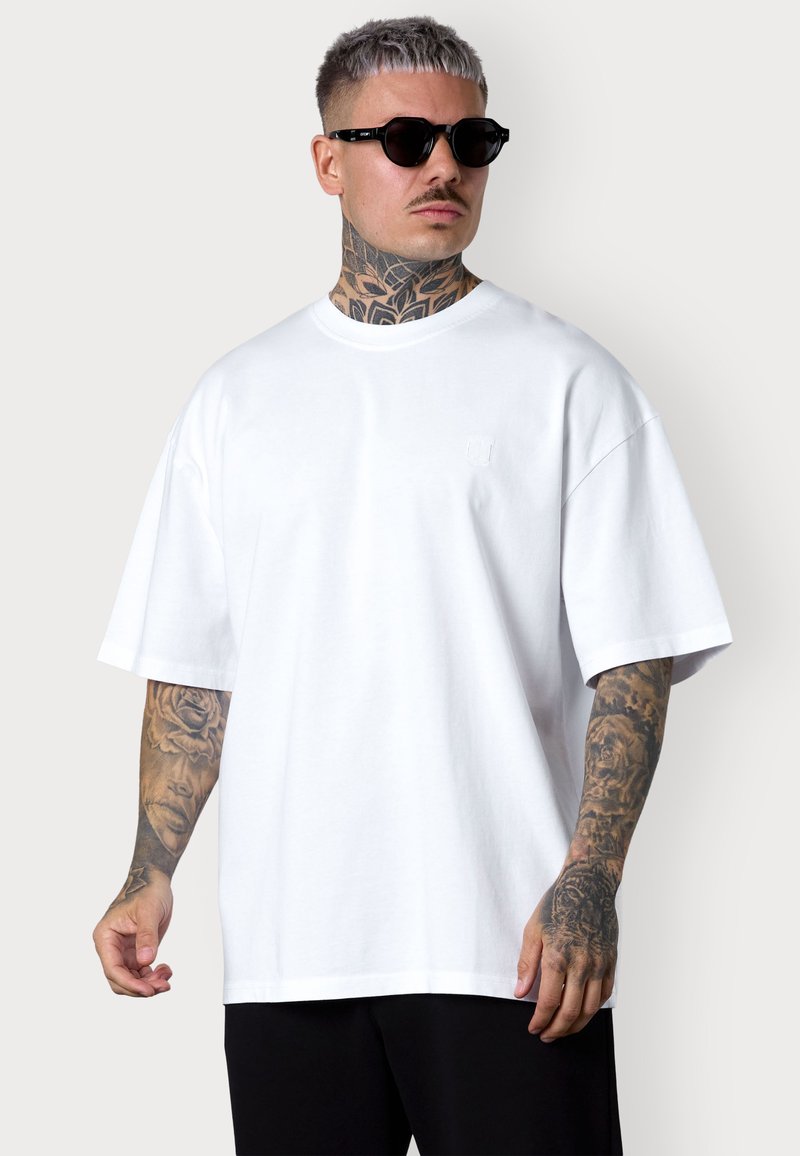 White oversized t-shirt with a crew neckline and short sleeves. Features subtle logo detail on the chest. Paired with black pants.