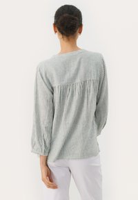 Bluză în dungi cu decolteu rotund, spate plisat și mâneci lungi cu manșete. Fabricată dintr-un material ușor în nuanțe de verde și alb.