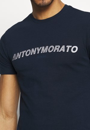 Camiseta de algodón navy con gran texto blanco/metálico "ANTONYMORATO" y el texto más pequeño "ITALIA" debajo. Cuello redondo, mangas cortas.
