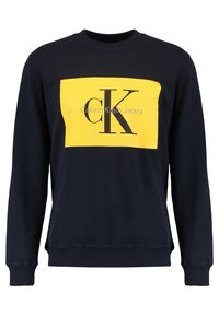 Sudadera azul marino con un panel rectangular amarillo que lleva el texto negro "Calvin Klein Jeans" y el logo "cK"; puños y dobladillo acanalados.