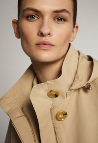 Beige waterdichte jas met een hoge kraag, voorzien van gele knopen en een ritssluiting. Glad textuur, traditionele ontwerp en geometrische stiksel.