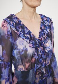 Vloeiende, doorschijnende jurk met een paars en blauw bloemenpatroon, geraffineerde halslijn en een gestrikte accenten bij de borst. Lange, vloeiende mouwen.