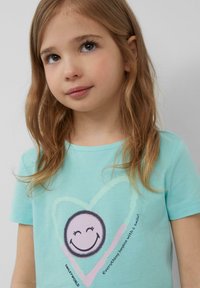 Turquoise katoenen t-shirt met korte mouwen, voorzien van een centrale smiley-graphic omringd door een hartomtrek en de tekst "Alles begint met een glimlach!"