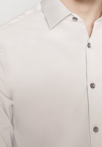 Camisa de botones gris claro con una textura suave, que presenta un cuello en contraste y botones oscuros a lo largo de la tapeta.