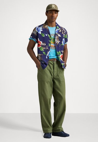 Camisa de manga corta con estampado floral en tonos azules, verdes y rojos, combinada con una camiseta gráfica turquesa y pantalones cargo verde oliva. Zapatos azul marino.