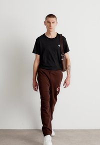 Melna kreklu ar apaļu kaklu, brūnas sporta bikses ar sarkanu logo un baltas tenisa kurpes. Brūns soma ar regulējamu siksniņu. Neitrāls fons.