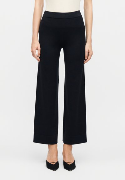 Femme portant un pantalon noir à jambes larges et des chaussures noires à bout pointu, debout devant un fond blanc uni.