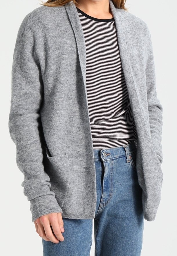 Personne portant un cardigan gris ouvert sur un chemisier rayé noir et blanc avec un jean bleu, debout devant un fond uni.