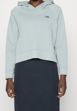 Person trägt einen hellblauen Kapuzenpullover mit kleinem Logo und einen dunklen, gesteppter Rock.