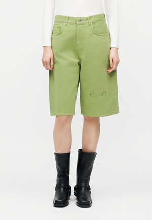 Osoba ubrana w jasnografitowe krótkie spodenki sięgające do kolan z przednimi kieszeniami i haftem "STIEGLITZ" na lewej nogawce, w połączeniu z czarnymi butami za połowę łydki.