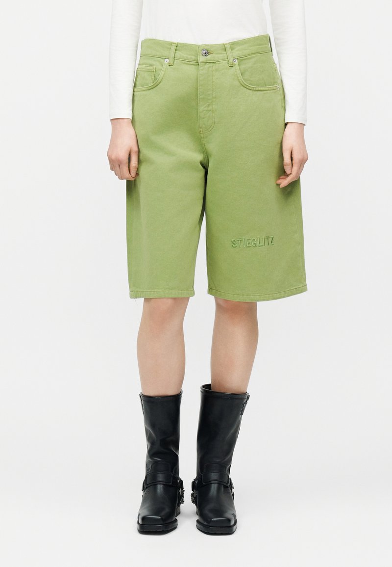 Isik, kes kannab helerohelisi põlveni ulatuvaid lühikesi pükse, millel on esiküljel taskud ja vasaku jala küljes tikandiga "STIEGLITZ", kombineerituna mustade kesk-sääre saapadega.