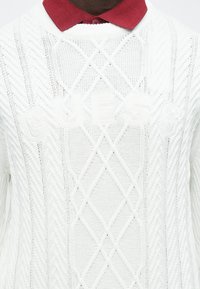 Osoba ubrana w biały sweter z grubym splotem z napisem „GUESS” oraz czerwoną koszulę z kołnierzykiem pod spodem, widoczna od ramion do połowy tułowia.