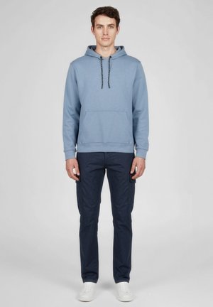 Man staat rechtop en kijkt naar voren. Hij draagt een lichtblauwe hoodie, donkerblauwe cargobroek en witte sneakers tegen een effen grijze achtergrond.