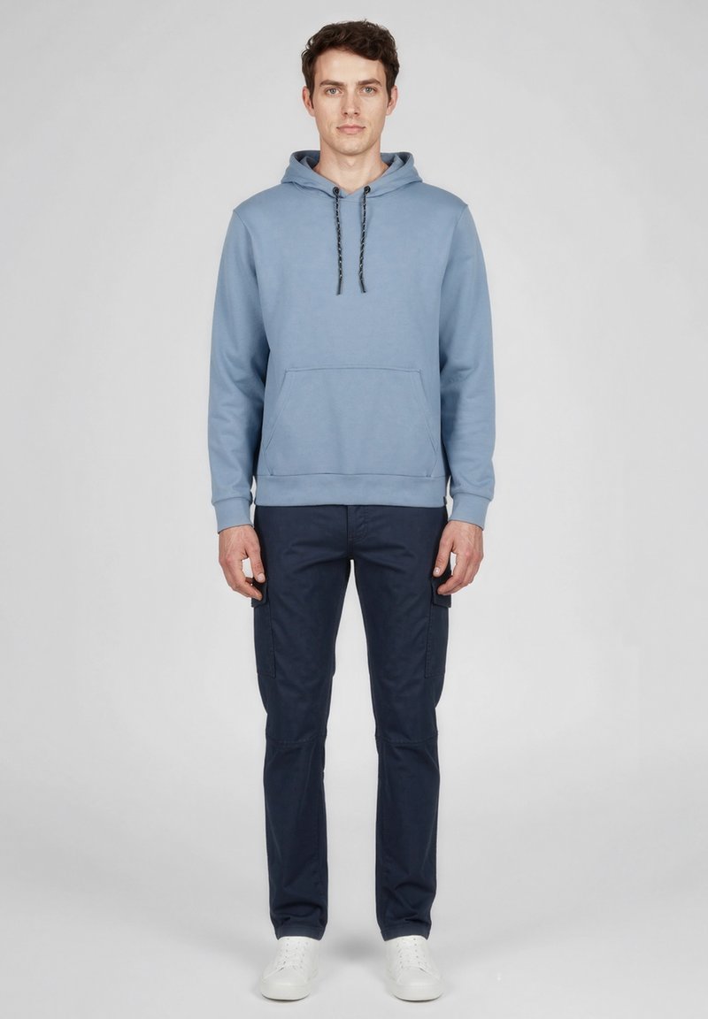Homme debout face à l'avant portant un sweat à capuche bleu clair, un pantalon cargo bleu foncé et des baskets blanches sur un fond gris uni.