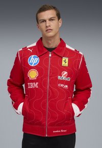 Giovane uomo che indossa una giacca rossa Scuderia Ferrari con loghi degli sponsor tra cui HP, Shell, IBM, UniCredit e Ceva Logistics, mani nelle tasche.