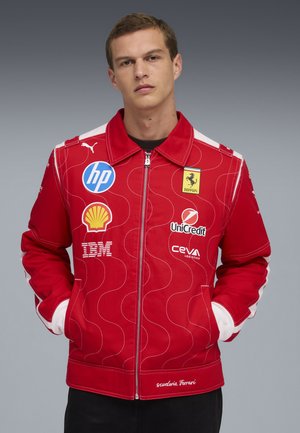 Joven con una chaqueta roja de Scuderia Ferrari, que lleva logotipos de patrocinadores como HP, Shell, IBM, UniCredit y Ceva Logistics, con las manos en los bolsillos.